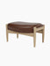 A side-profile view of The Seal Ottoman, showing its plush brown leather cushion and natural oak frame with tapered legs.