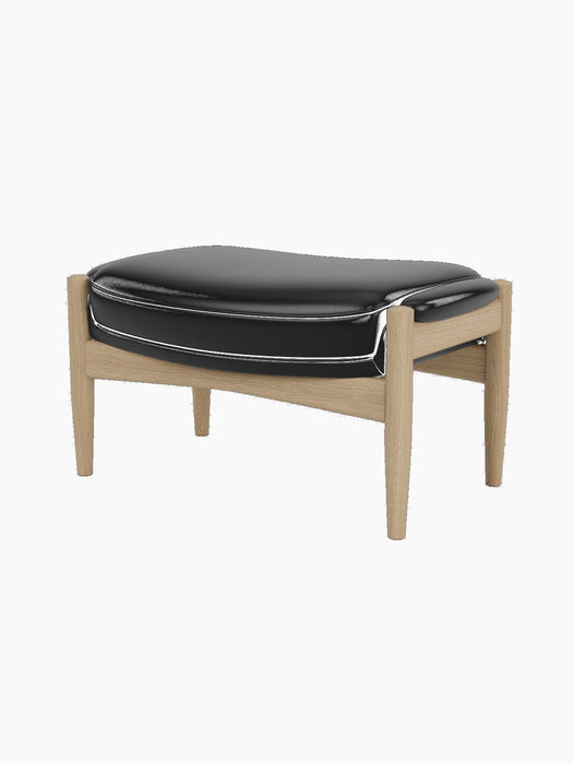 The Seal Ottoman, shown at an angle, features a plush black leather cushion resting on a sculpted natural oak frame.