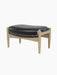 The Seal Ottoman, shown at an angle, features a plush black leather cushion resting on a sculpted natural oak frame.