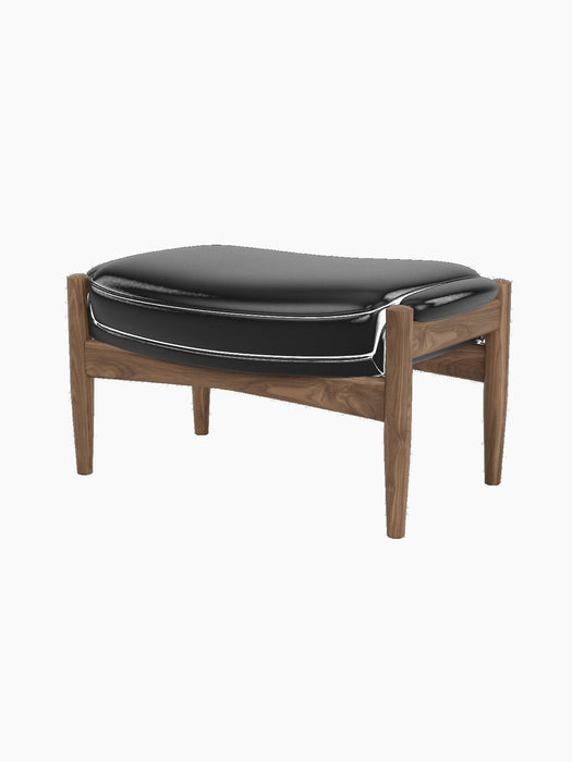 A three-quarter view of The Seal Ottoman, featuring a solid walnut frame with curved supports and a padded black leather top.