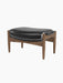 A three-quarter view of The Seal Ottoman, featuring a solid walnut frame with curved supports and a padded black leather top.