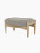 A side view of The Seal Ottoman, featuring a light grey upholstered cushion on a mid-century modern frame of natural oak.