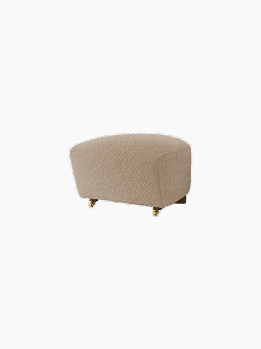 The Tired Man Ottoman in beige textile with dark wood legs and front brass casters, pictured on a black background.