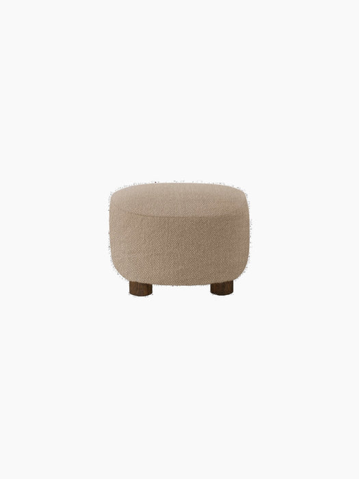 A back view of The Tired Man Ottoman, upholstered in a light brown textile, standing on four short, dark-stained oak legs.