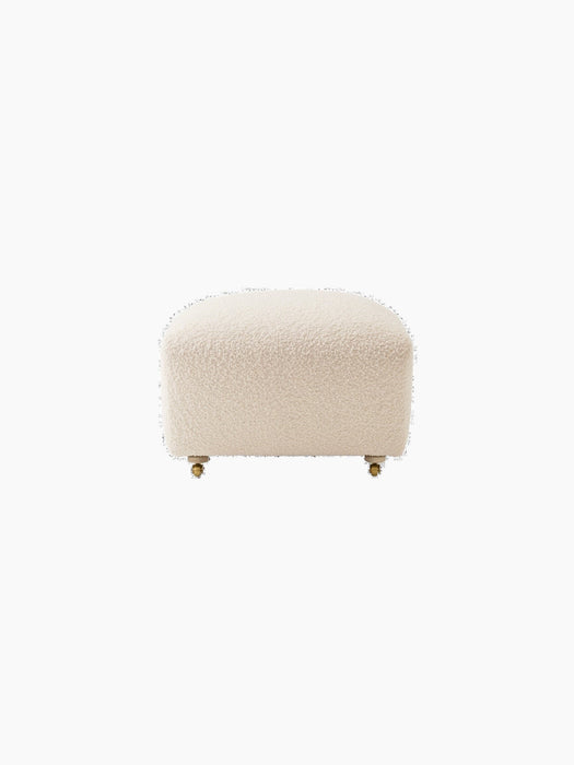 A front view of The Tired Man Ottoman, upholstered in a plush, off-white textile, with light oak legs and brass casters.