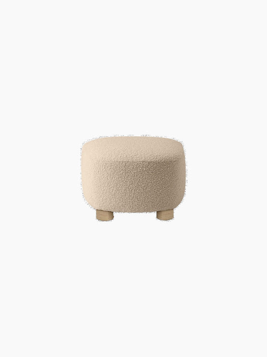The Tired Man Ottoman, seen from the back, upholstered in a cream textured fabric with four short, stout light oak legs.