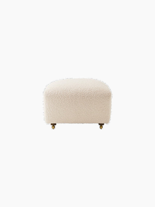 Front view of The Tired Man Ottoman upholstered in cream-coloured textured textile, with short legs and small brass casters.