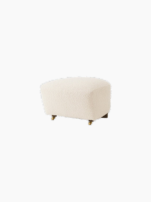 The Tired Man Ottoman in cream bouclé fabric, shown at an angle. It has dark wood legs with two brass casters at the front.