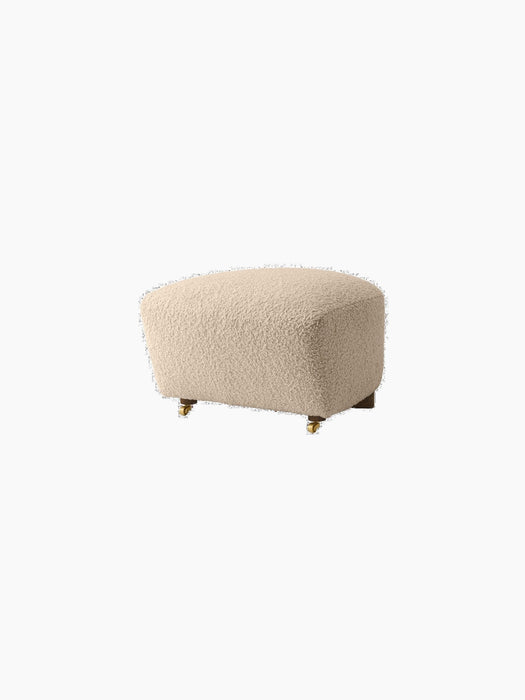The Tired Man Ottoman in a textured cream fabric, shown at an angle, with dark wooden legs and small brass castors.