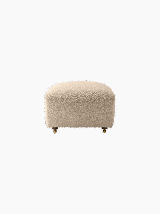 A front view of The Tired Man Ottoman, featuring its plush, off-white textured upholstery and short legs with brass casters.