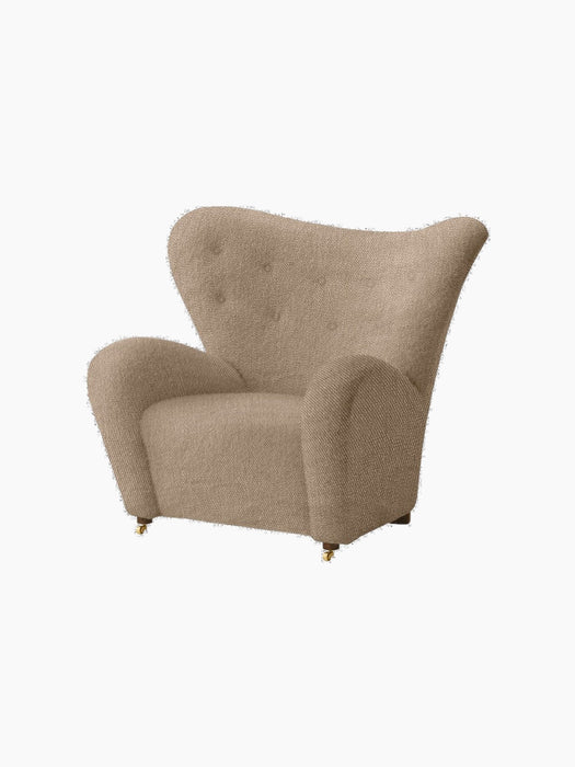 Angled view of The Tired Man Lounge Chair in light brown textile, featuring high curved back, plush arms, and brass castors