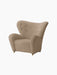Angled view of The Tired Man Lounge Chair in light brown textile, featuring high curved back, plush arms, and brass castors