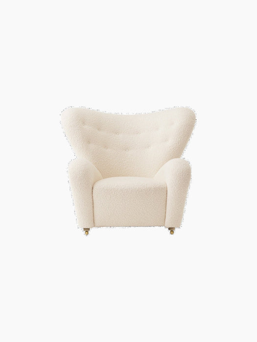 Front view of The Tired Man Lounge Chair in cream textile with a high, curved back, rounded arms and brass castors.