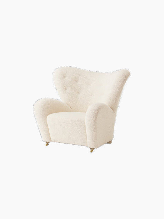 An angled view of The Tired Man Lounge Chair in a textured off-white textile, showing its curved high back and brass castors.