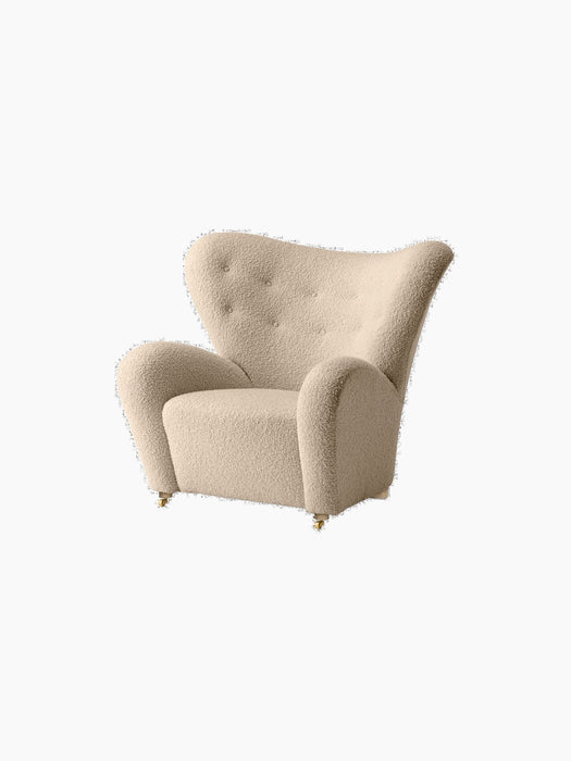 The Tired Man Lounge Chair upholstered in a cream textile, featuring a high curved back, tufted details, and gold castors.