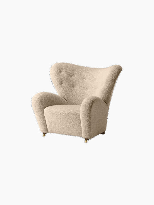 Angled view of The Tired Man Lounge Chair in plush, cream-coloured textile with a high, tufted back & brass front castors.