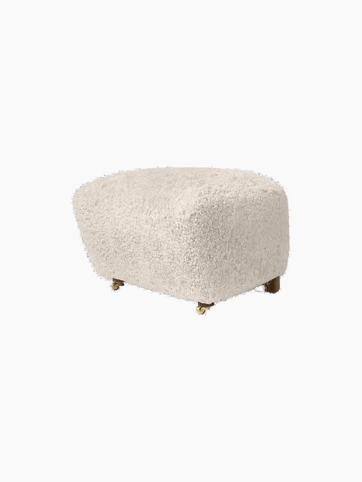 The Tired Man Ottoman, upholstered in plush Moonlight sheepskin with dark stained oak legs and small brass front wheels.