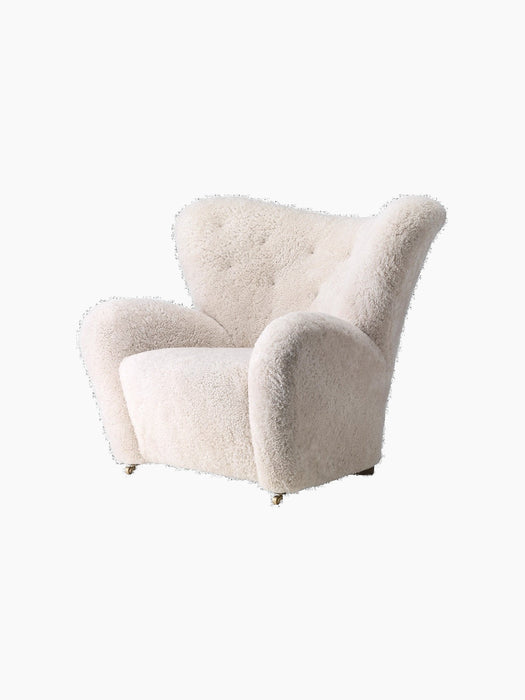 Side view of The Tired Man Lounge Chair in off-white sheepskin, showing its curved high back, large arms, and brass wheels.
