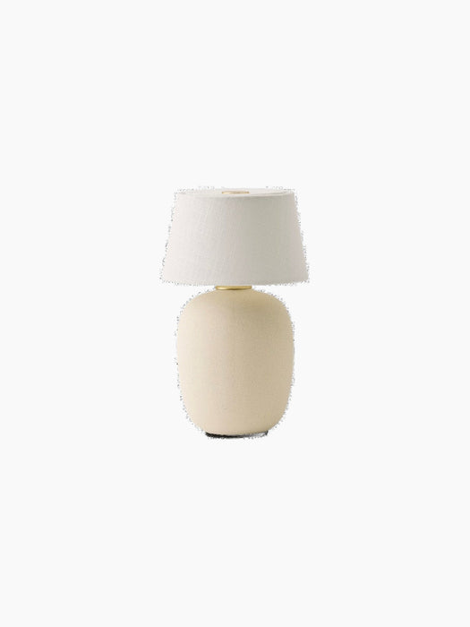 A Torso Portable Table Lamp in Sand, with a rounded, textured ceramic base and a white linen shade on a black background.