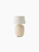 A Torso Portable Table Lamp in Sand, with a rounded, textured ceramic base and a white linen shade on a black background.
