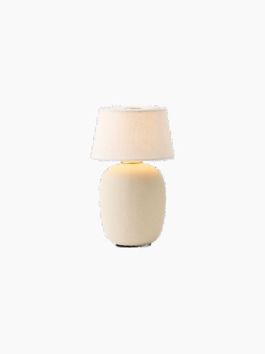 The illuminated Torso Portable Table Lamp with a sand ceramic base and off-white linen shade, topped with a brass button.