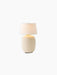 The illuminated Torso Portable Table Lamp with a sand ceramic base and off-white linen shade, topped with a brass button.