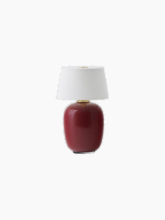 The Torso Portable Table Lamp in Ruby, with a glossy ceramic base, a white linen lampshade, and a brass touch-button on top.