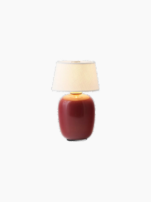 The Torso Portable Table Lamp in Ruby, illuminated against a black background, with a glossy ceramic base and linen shade.