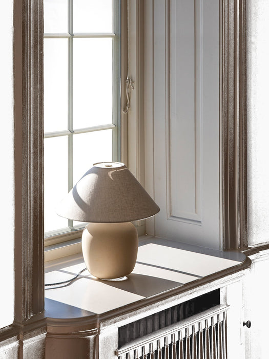 The Torso Table Lamp, 37, featuring a cream ceramic base and linen shade, is placed on a sunlit white windowsill.