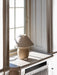The Torso Table Lamp, 37, featuring a cream ceramic base and linen shade, is placed on a sunlit white windowsill.