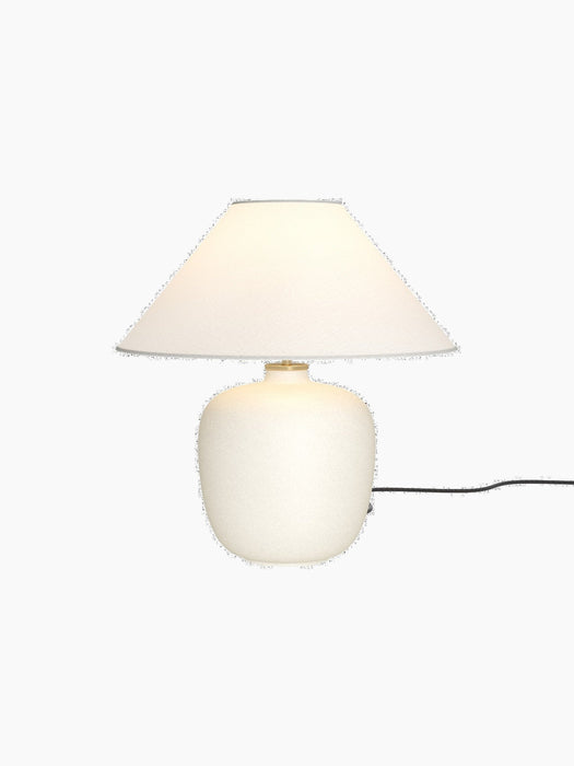 A front-on photograph of the Torso Table Lamp, 37. The lamp is lit, featuring an off-white ceramic base and a linen shade.