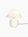 A front-on photograph of the Torso Table Lamp, 37. The lamp is lit, featuring an off-white ceramic base and a linen shade.