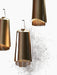 A close-up of three brass Umanoff Pendants hanging at different levels against a dark, ribbed wall, emitting a warm glow.