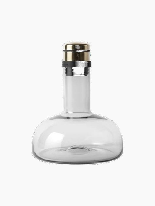 The Audo Copenhagen Wine Breather in Smoke-Brass. A clear glass decanter with a wide base and a brass and black stopper.
