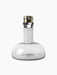 The Audo Copenhagen Wine Breather in Smoke-Brass. A clear glass decanter with a wide base and a brass and black stopper.